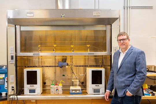 a man in a gray suit stands next to a lab in R I T college of engineering technology.