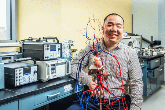 Smiling man in labratory holds a detailed anatomical model of a human heart with red and blue blood vessels. Electronic testing equipment and lab instruments are visible on the bench behind him.