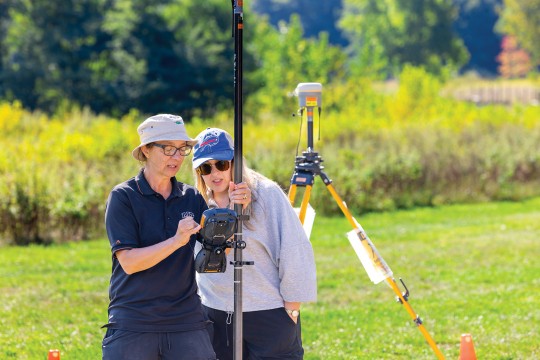 two women stand in a field using imaging equipment.