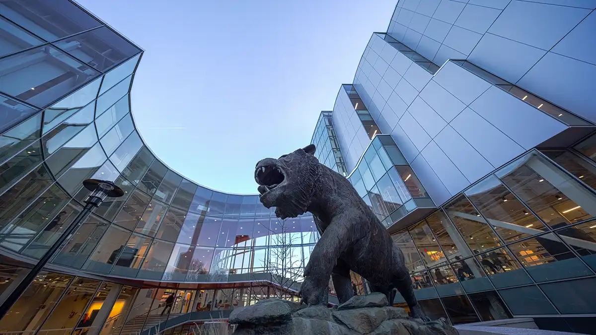 A bronze tiger statue stands roaring in front of a modern glass building on the R I T campus.