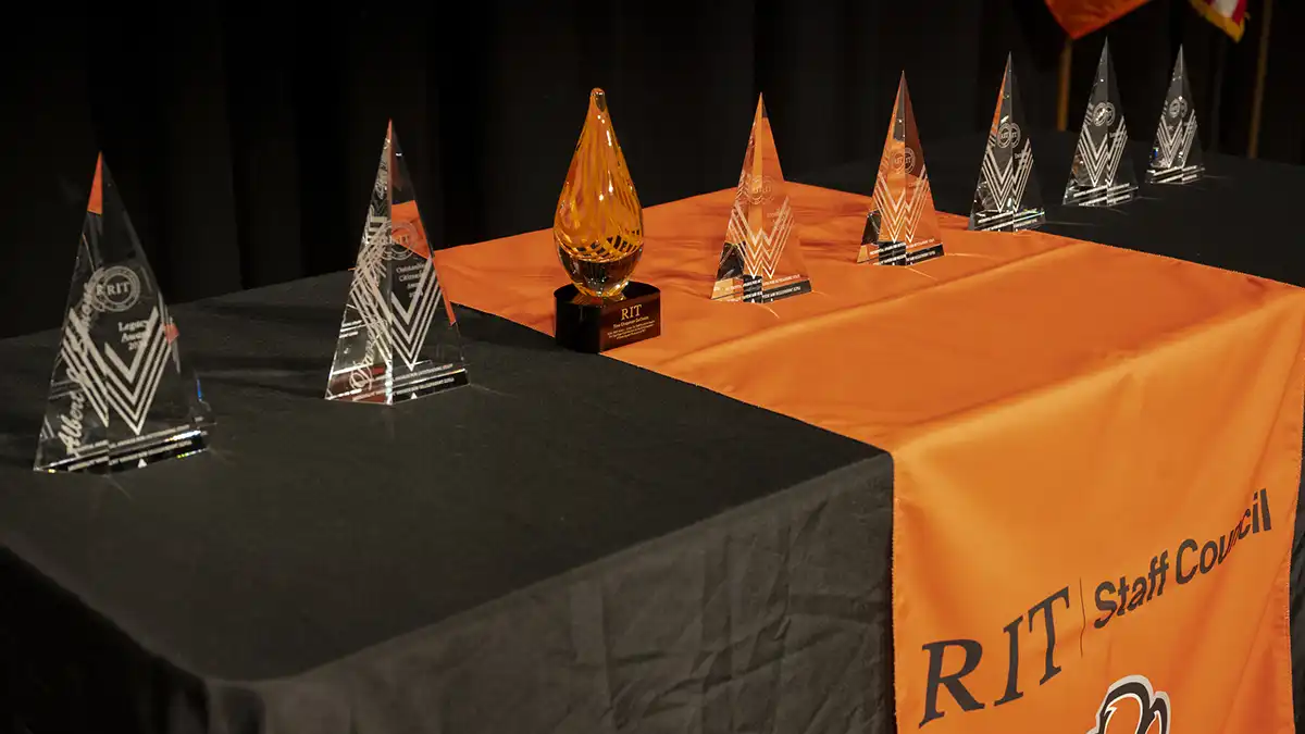 A row of triangular glass awards and one amber-colored award sit on a table draped with an R I T Staff Council cloth.