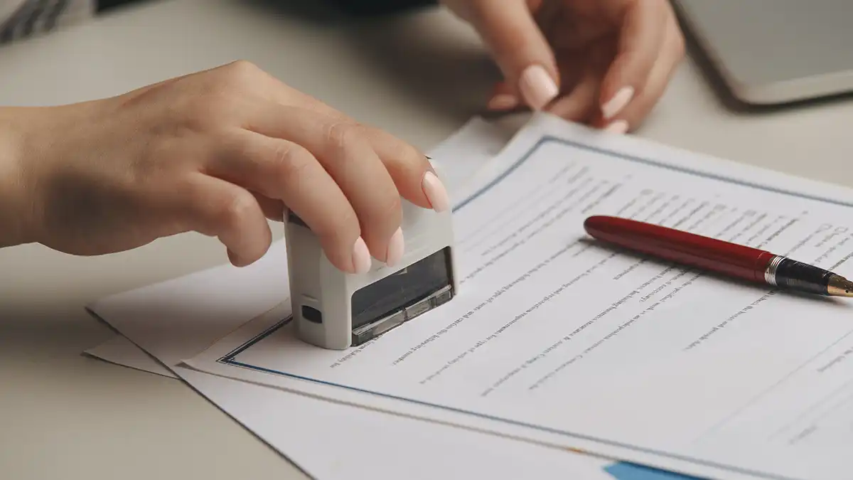 A close-up of a hand pressing a notary stamp onto an official document next to a red pen.