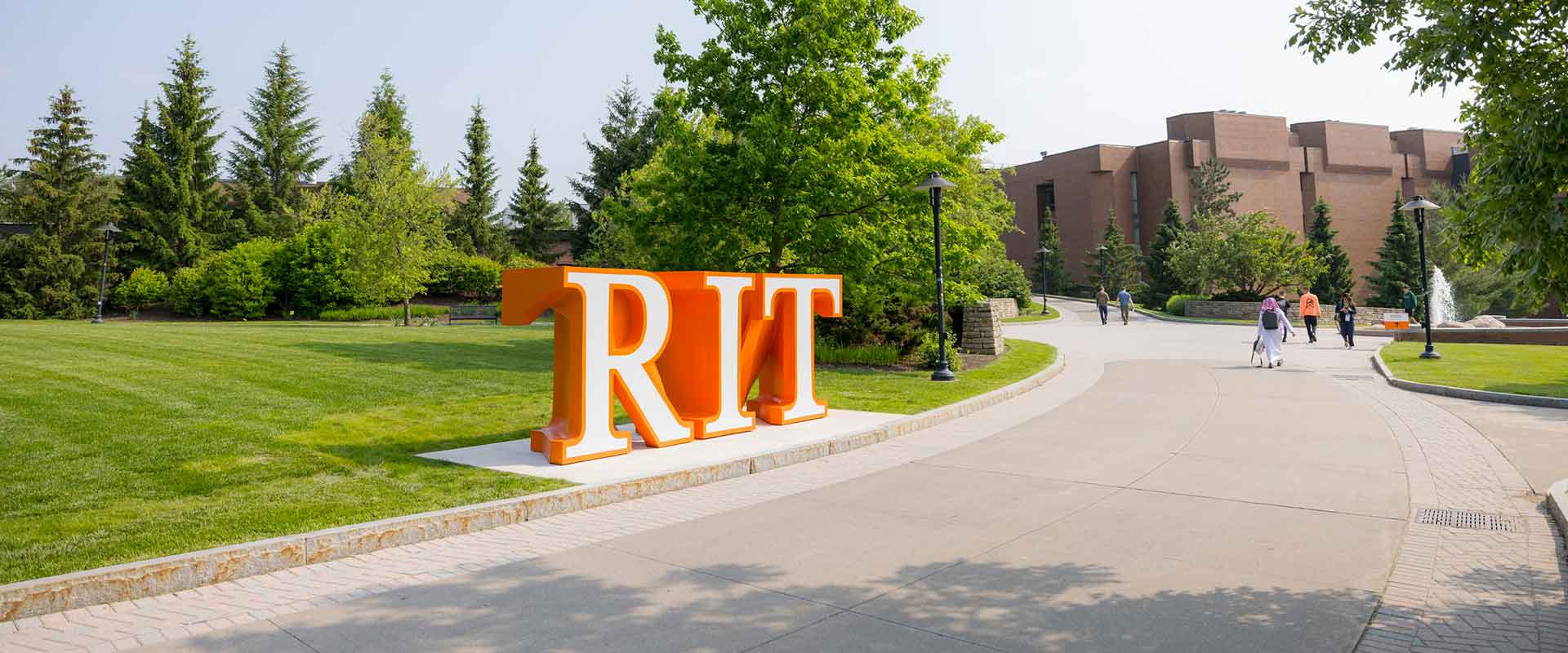 Large orange 'R I T' letters stand outdoors along a walkway surrounded by trees and campus buildings.