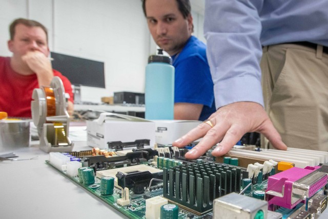 two adult student look on as a professor touches a large circuit board.