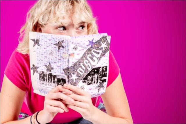 college student holding a small magazine up to her face as she peeks over the top of it.