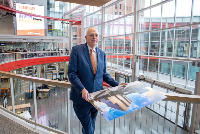 David Munson, Jr., RIT President, stands on the second floor balcony of the SHED holding a portrait that shows the renderings before it was built. The.