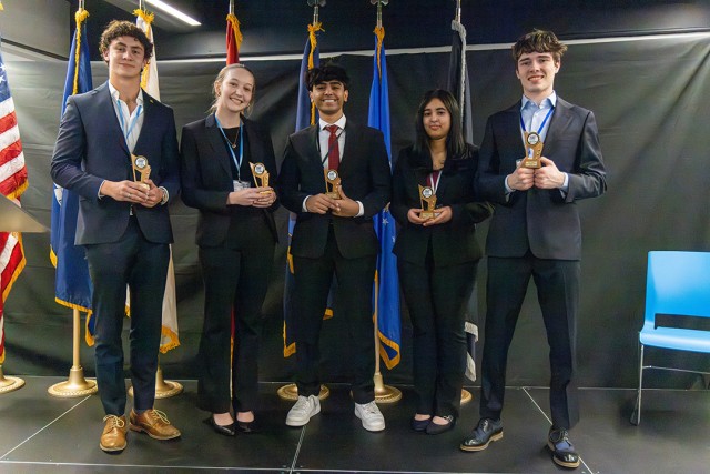 Five young professionals in formal attire stand on a stage holding small trophies, smiling. Behind them, multiple flags are displayed. The stage has a dark backdrop, and a blue chair is visible to the side.