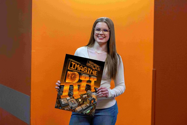 Smiling woman with long blonde hair and glasses holds a poster for 'Imagine RIT,' featuring a circuit board design with glowing orange text. She stands against an orange-toned background.