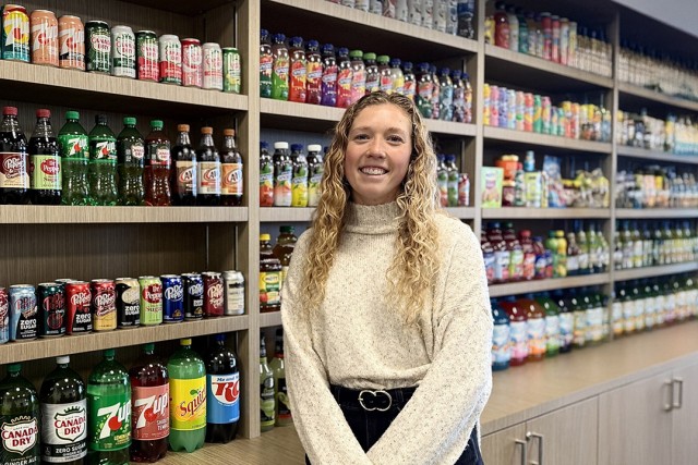 a woman in a cream sweater and black jeans stands in front of shelves displaying assorted sodas.