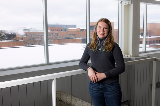 a young woman dressed in jeans and a black sweater looks at the camera and leans on a railing next to windows looking out on to a snow covered campus.