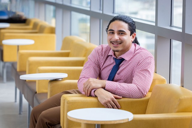 Person smiling while seated in a modern lounge area with mustard chairs and large windows.