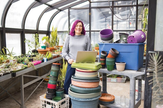 a woman stands in a greenhouse at R I T with plants and planters of various sizes surrounding her.