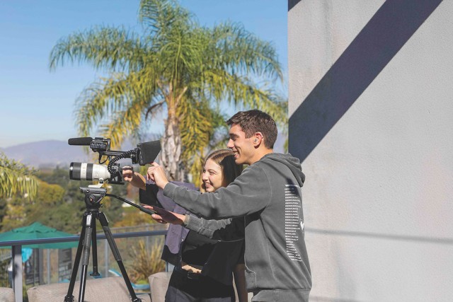 Two people operating a video camera on a terrace with a palm tree and hills in the background.