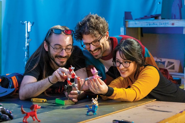 three adults huddle close together holding figurines used to make a stop motion animated film in an art studio setting.