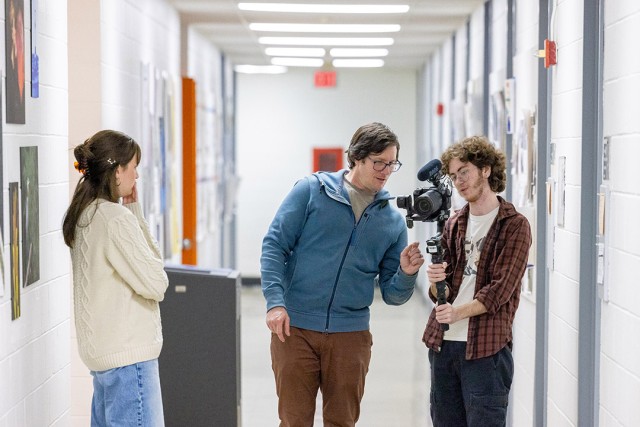 An AP Video Journalist shows some camera handling techniques to two students in a college building hallway.