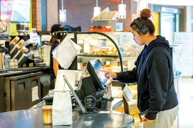 a student in a black hoodie and headphones leans over a countertop kiosk in a cafe entering information.