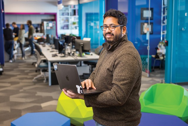 a man in a brown sweater stands in a bright multicolored room holding a laptop.
