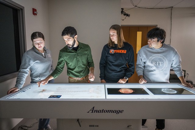 A priofessor and three students stand around a digital cadaver table, which allows for interactive, 3D exploration of the human body.
