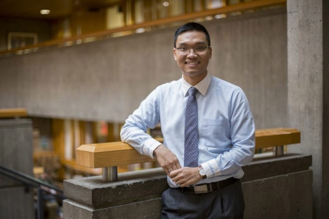 a man in a light blue shirt and tie leans against a wooden railing in an open room with gray walls.