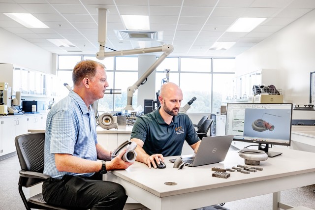 Two men in a lab reviewing a 3D mechanical part design on a computer.