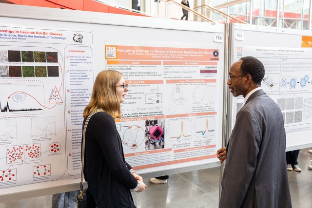 Female student in glasses explains a radiative-efficiency poster to a male researcher.