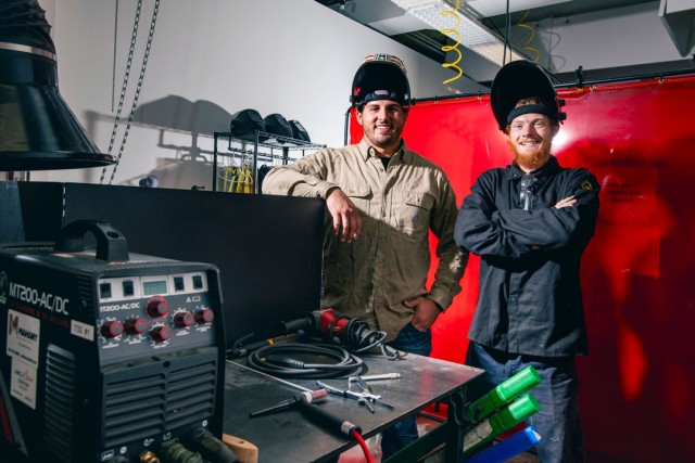 Two people wearing welding helmets stand behind a welding bench with equipment.