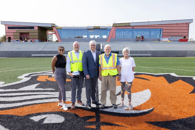 Five people standing on a sports field with a construction site in the background.
