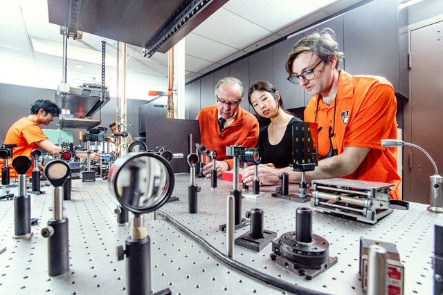 A group of people in orange and black tops work in a lab.