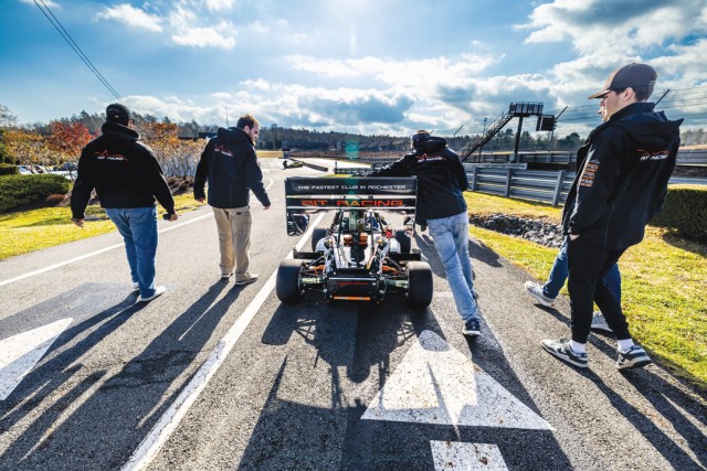 students walk forward next to a an R I T Formula S A E car on a track wearing black jackets.