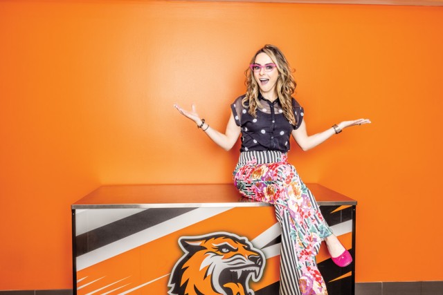a woman with long brown hair sits on an RIT podium in brightly colored clothing and pink glasses.