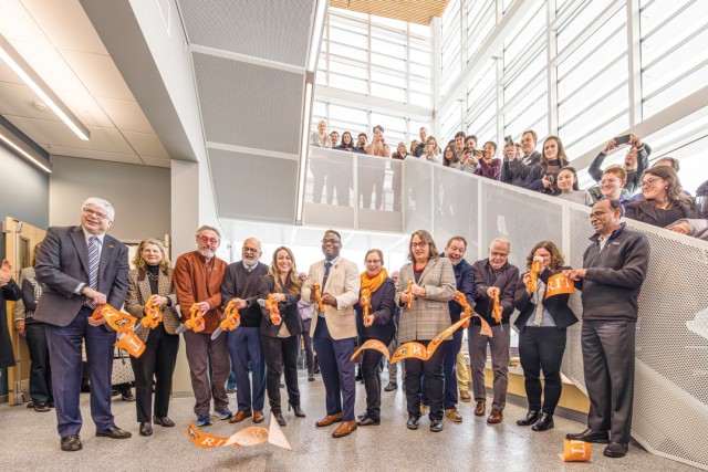 a group of people stand in a modern building cutting a large orange R I T ribbon with large scissors.