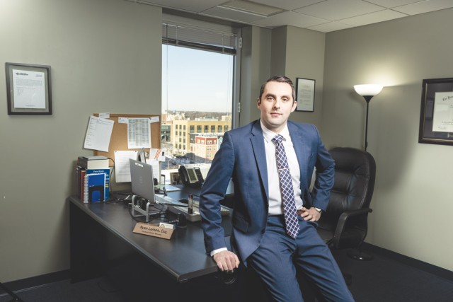 a man in a blue shirt leans on a desk in an office with a window looking out in to a city view.