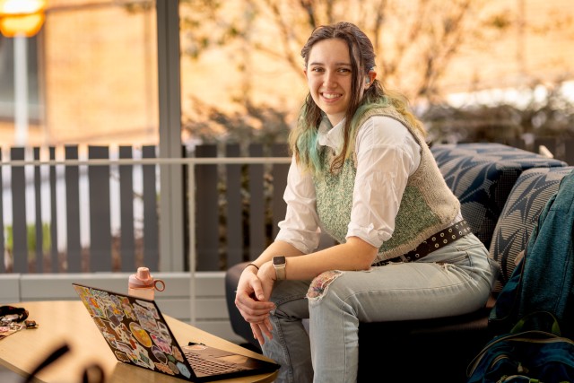 a college age woman with long brown hair sits on a porch in front of a laptop.