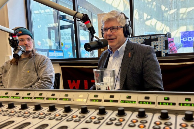 a student sits with a man in a gray suit inside a radio studio speaking in to a hanging microphone