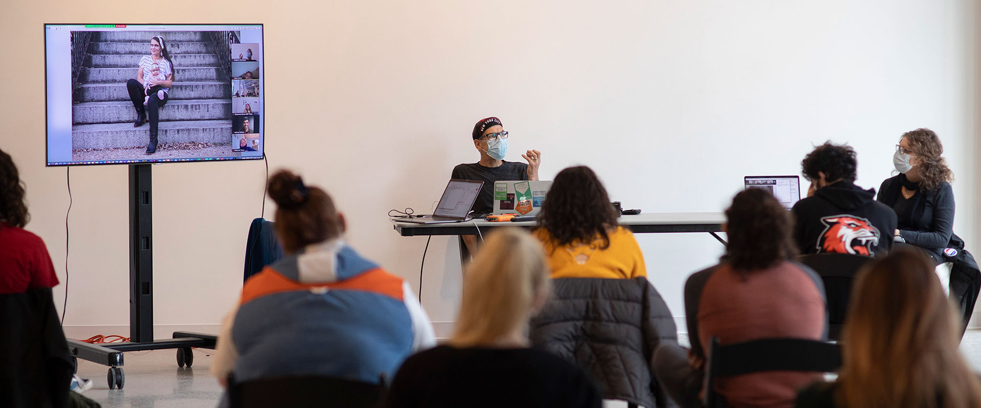 a group of seated audience members listening to a presenter seated at the front of the room, next to a T V screen showing a person sitting on steps outside.