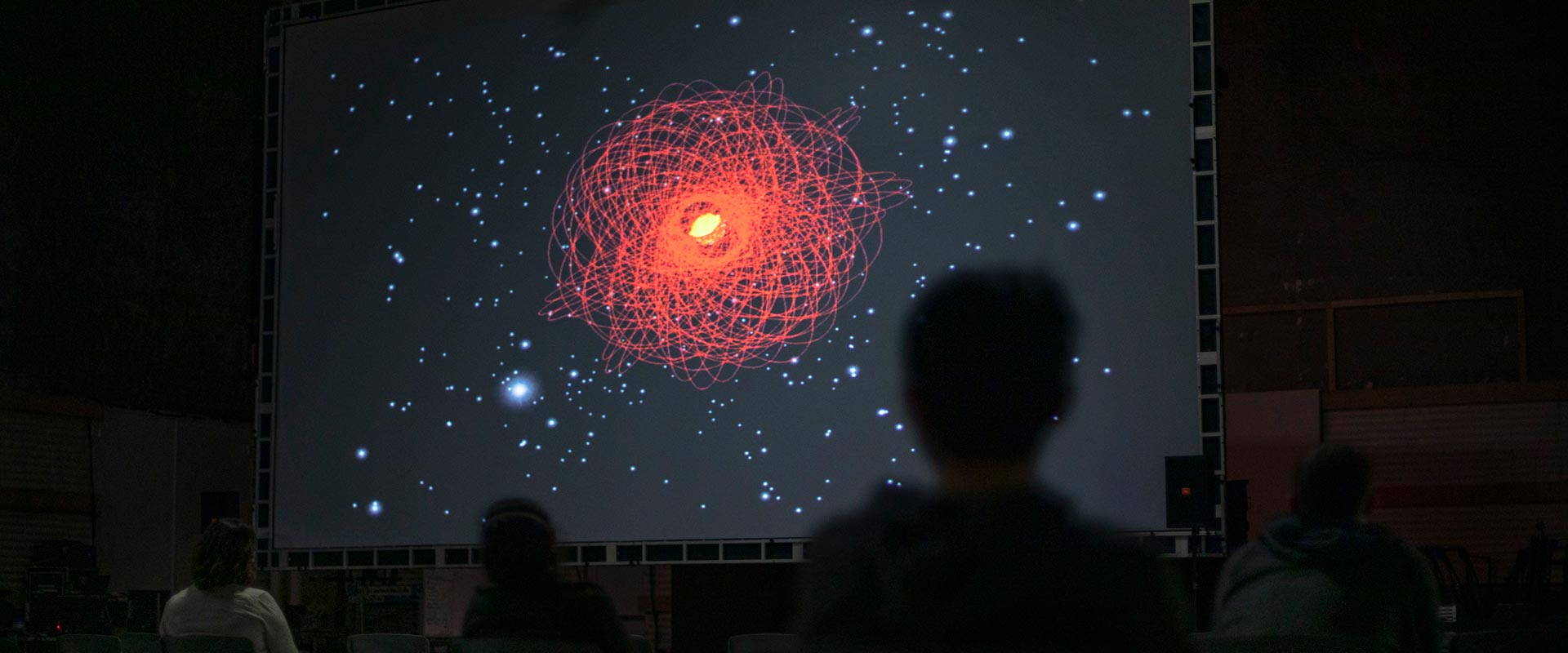 audience members sit in a dark room watching a screen showing a burst of red light with red lines swirling around it.