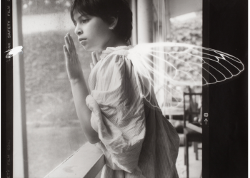 Black and white photograph of young girl looking out window with cicada wings on her back