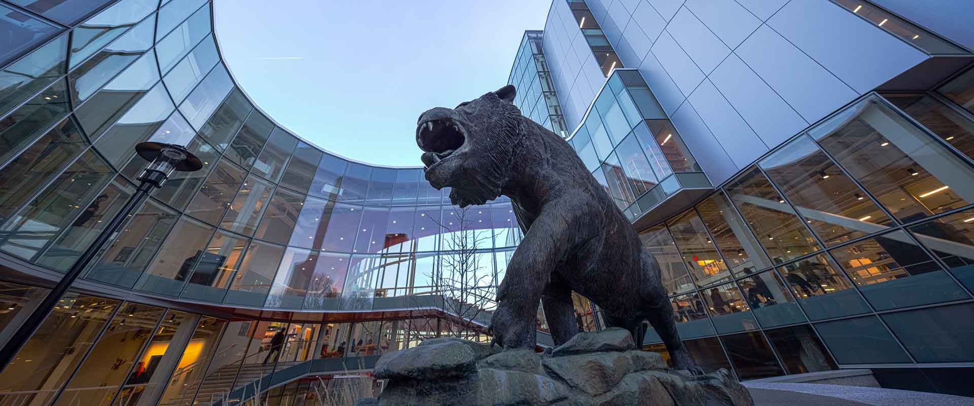 Low-angle view of the RIT tiger statue in a modern glass courtyard with surrounding buildings.
