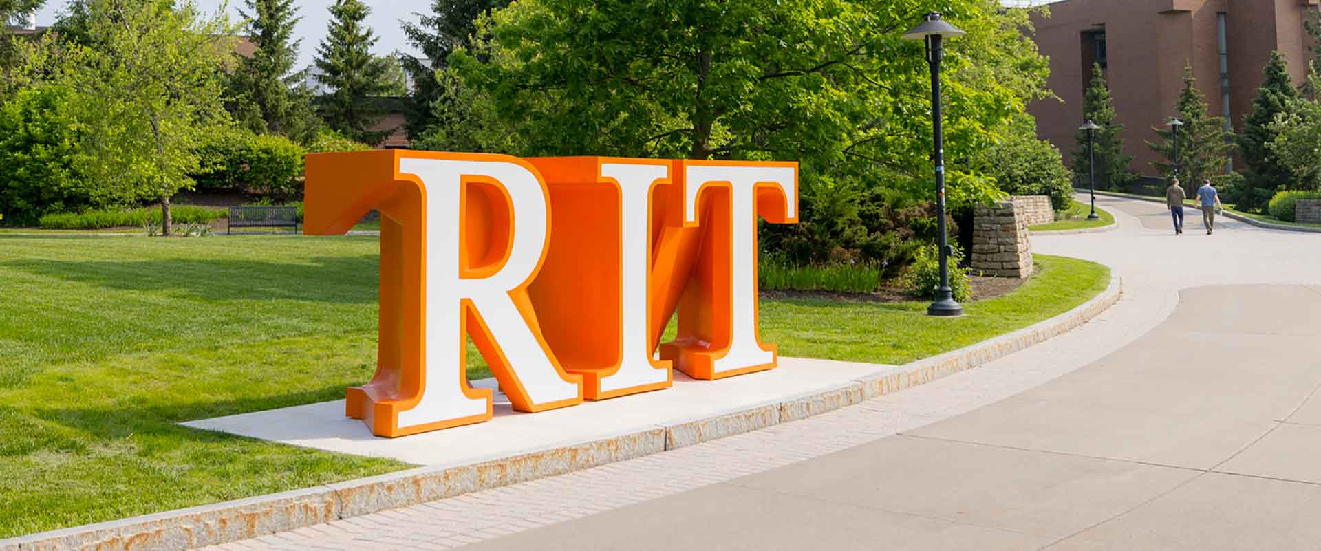 Large orange and white RIT letters on a landscaped path with trees and people in the background.
