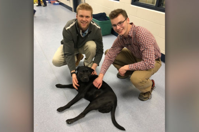 Sean and Will pet a medium-sized black puppy who is laying on the floor. The puppy is missing one of her back legs.
