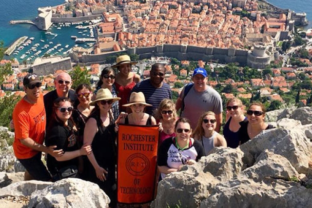 A group of RIT students and parents pose on the mountainside in Croatia, a beautiful city landscape behind them. They hold a banner that reads "Rochester Institute of Technology."