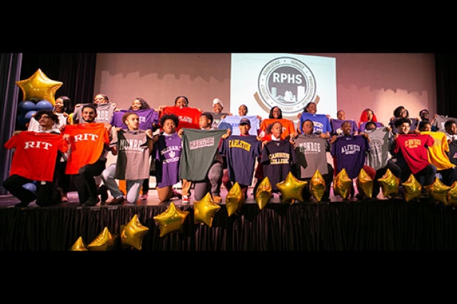 Students on stage holding up their future colleges' shirts.