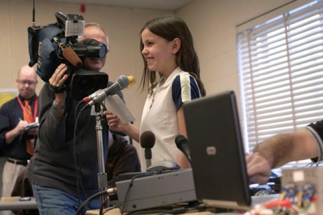 A young person speaks at a microphone while being filmed.