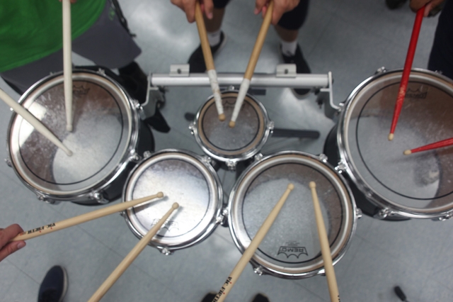A photo looking down on a drum set with five people holding out their drum sticks over the drums, as if they are about to all play the drums at once.