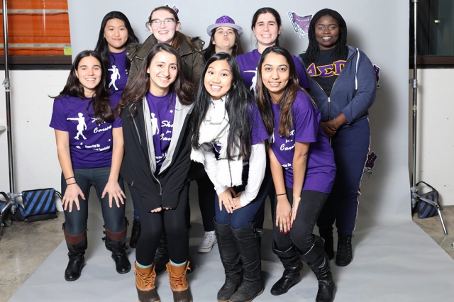Group of nine female students wearing purple T-shirts stands together in front of white backdrop