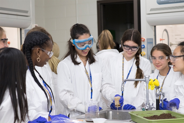 Girls in lab coats wearing safety goggles conducting an experiment over a sink.