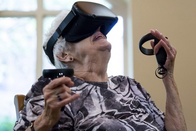 Elderly woman uses virtual reality goggles and remotes.