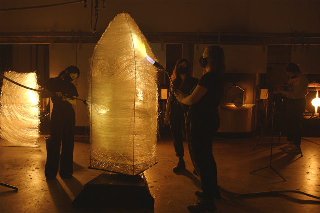 A group of artist take flame torches to a large, woven sculpture.