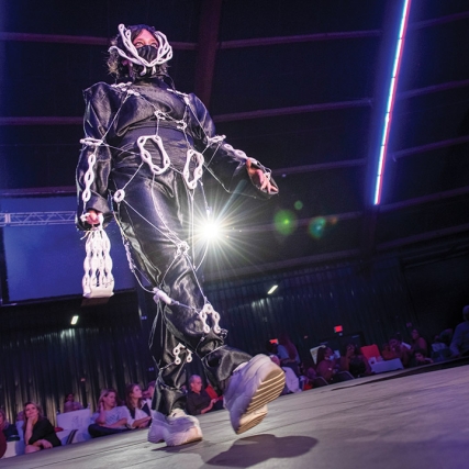student walking a runway wearing a piece that features black fabric and white chain-like structures.