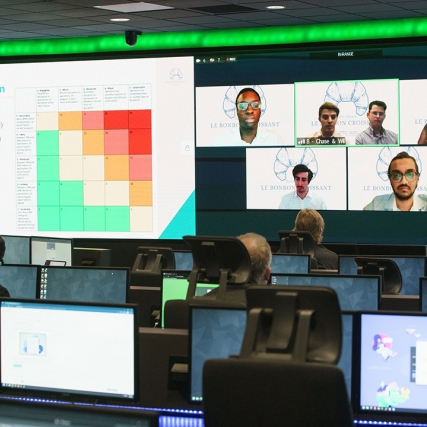 students sitting at rows of computers with several large screens projecting charts and team members' faces.